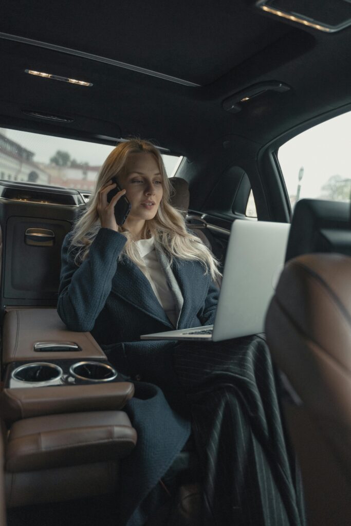 A woman traveling for business in a car talking on her cell and working on her laptop.